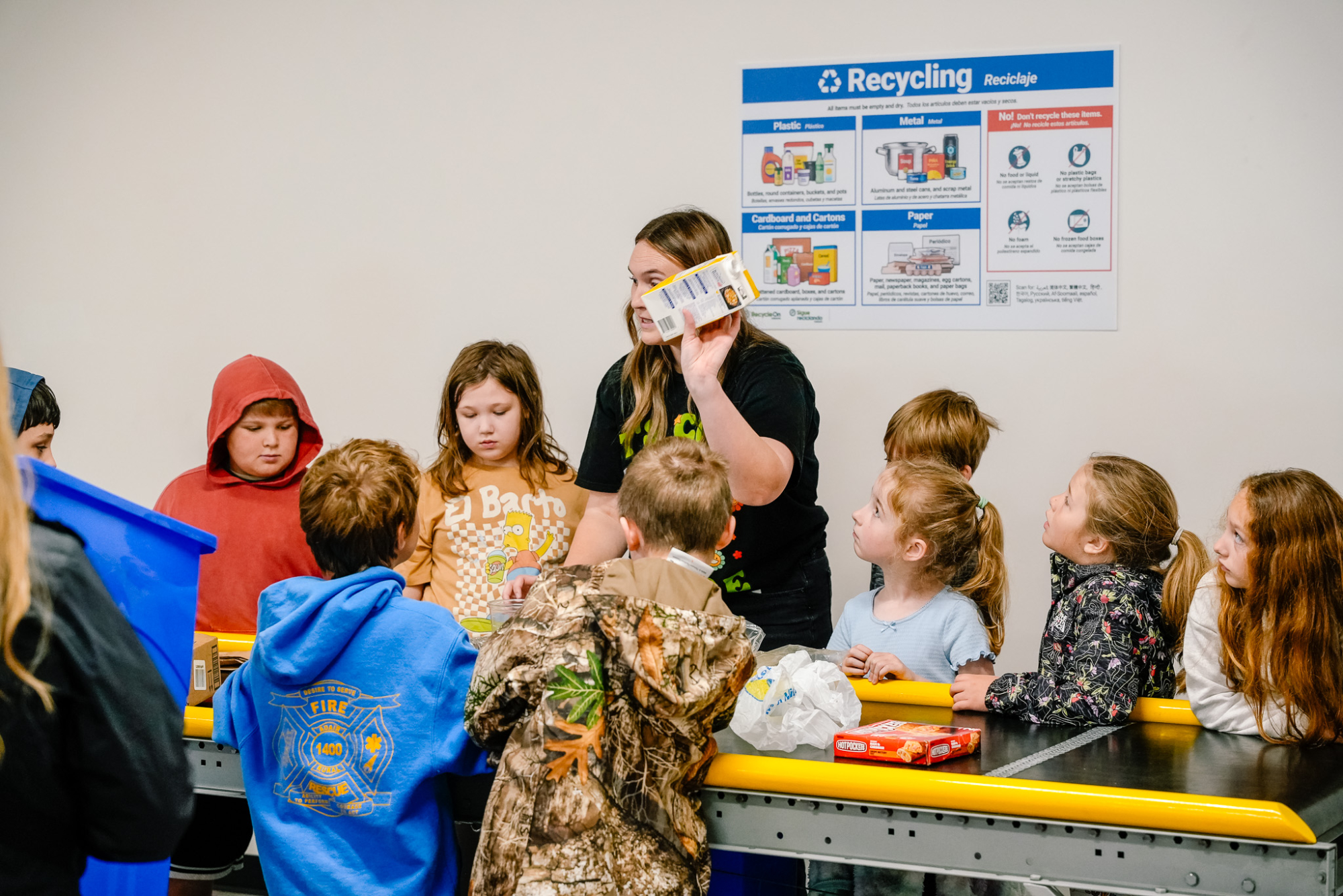 A group of children are gathered around a toy conveyor belt looking up at a woman with long hair holding an empty soup carton as she teaches them how to sort waste and recycling.